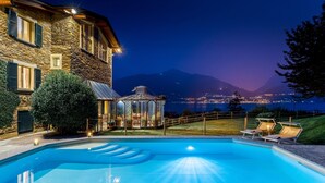 Un bâtiment en pierre doté d’une piscine et d’un belvédère, offrant une vue sur un lac et des montagnes la nuit.