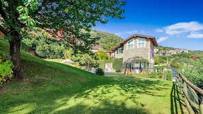 Charming stone house nestled in a lush hillside setting with a well-maintained lawn and a variety of trees.