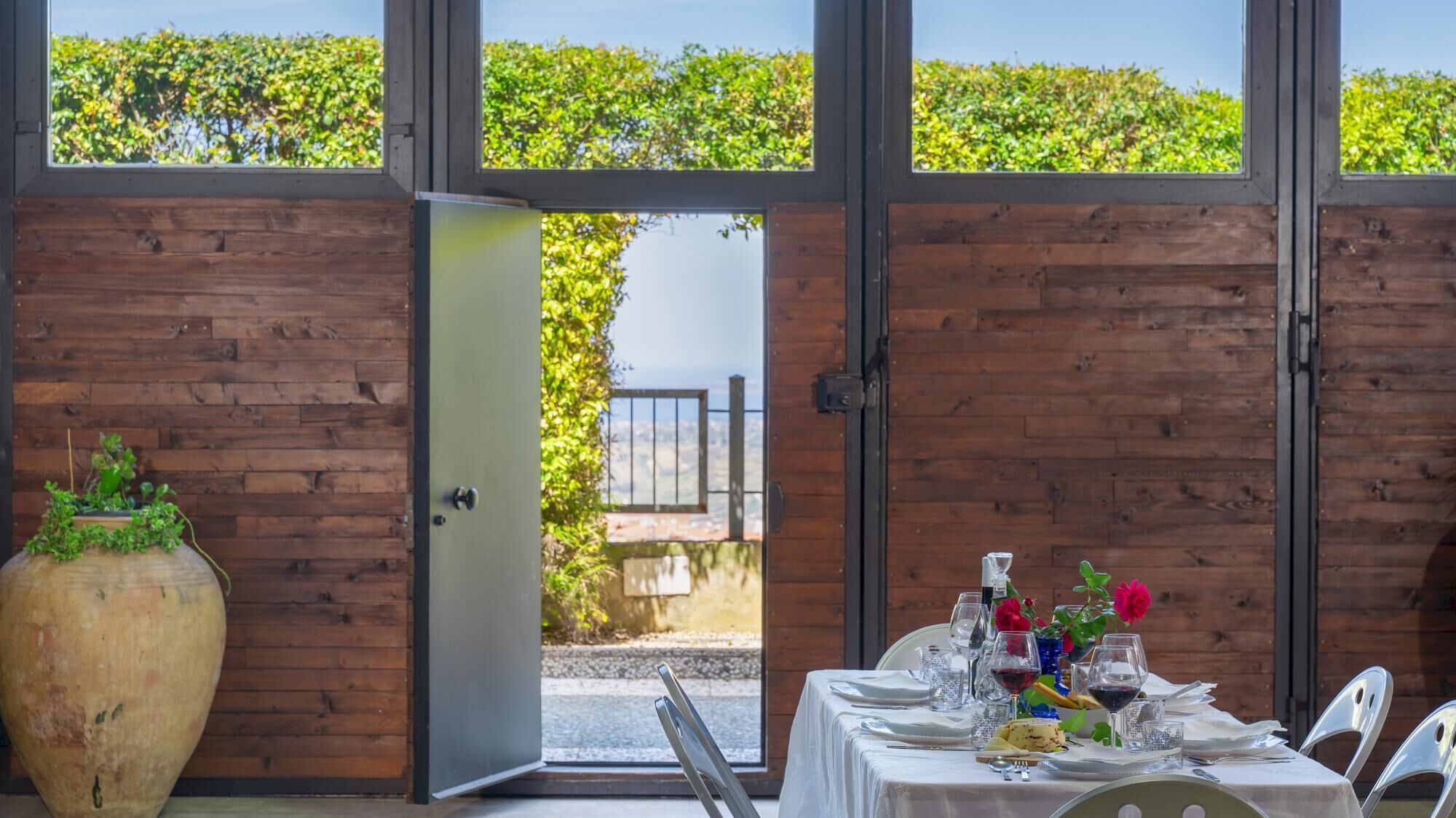 A dining area with a table set for two, a large vase with greenery, and a view of a wooden fence and greenery outside.