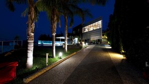 Un bâtiment moderne, avec un chemin menant à l’entrée, entouré de palmiers et doté d’espaces extérieurs bien éclairés.