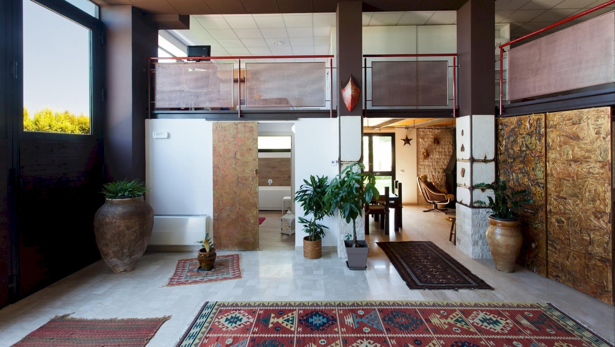 A spacious lobby with a high ceiling, large windows, and a view of greenery outside. There are several potted plants and a large rug on the floor.