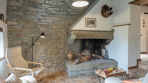 A cozy living room with a stone wall, a fireplace, a comfortable chair, and a table with a basket and glass. - Casa Largnano 6 2 in Arezzo (Castiglion Fiorentino)