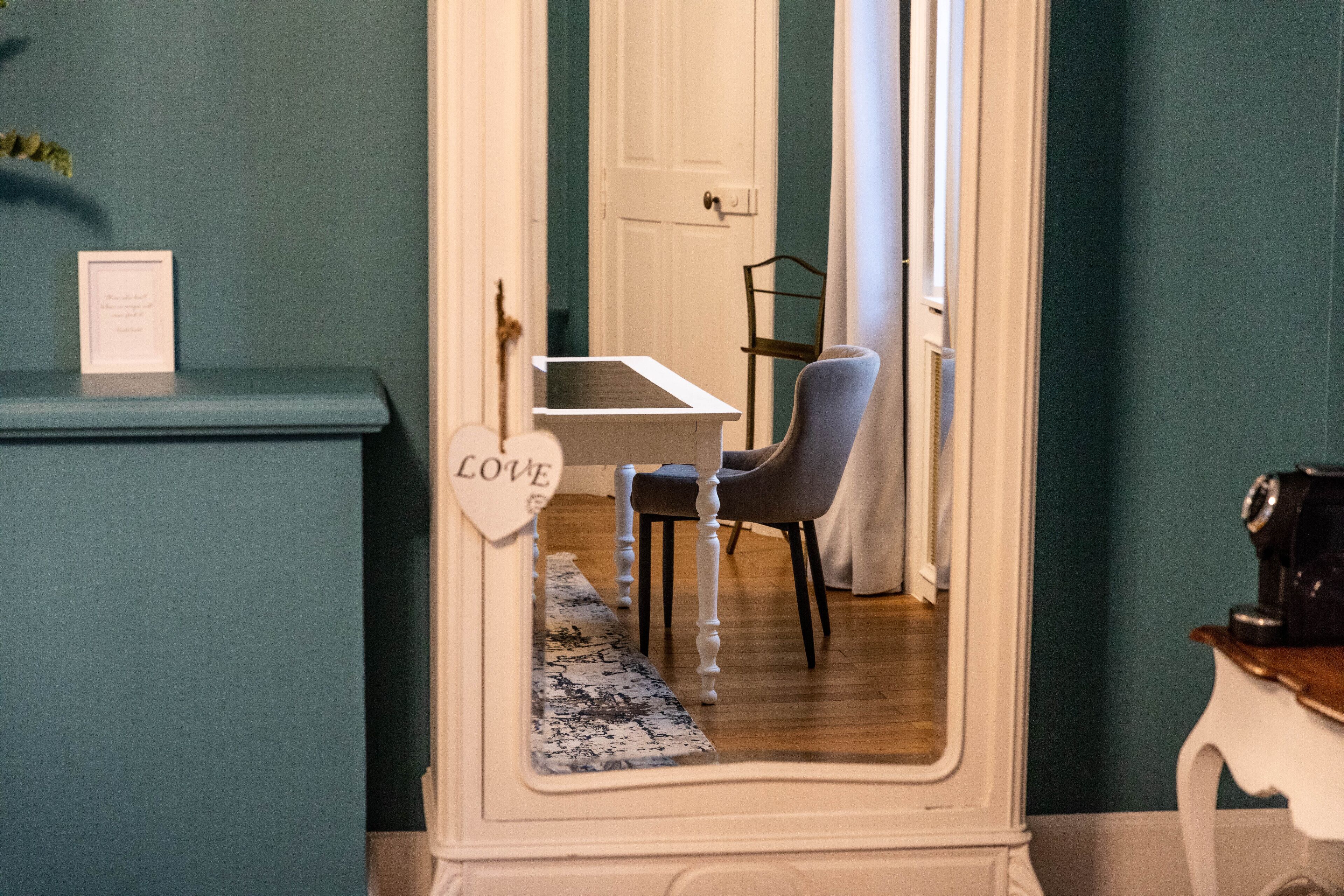 A room with teal walls, a white door, a small table with a chair, and a framed picture on a blue and white patterned rug.
