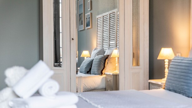 A neatly made bed with pillows and a rolled-up towel on the side, a bedside table with a lamp, and a mirror reflecting the room's interior. - Ô Château (Hayange)