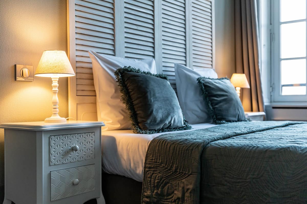 Una cama bien hecha con un edredón verde oscuro, dos lámparas de noche y una mesita de noche con cajones.