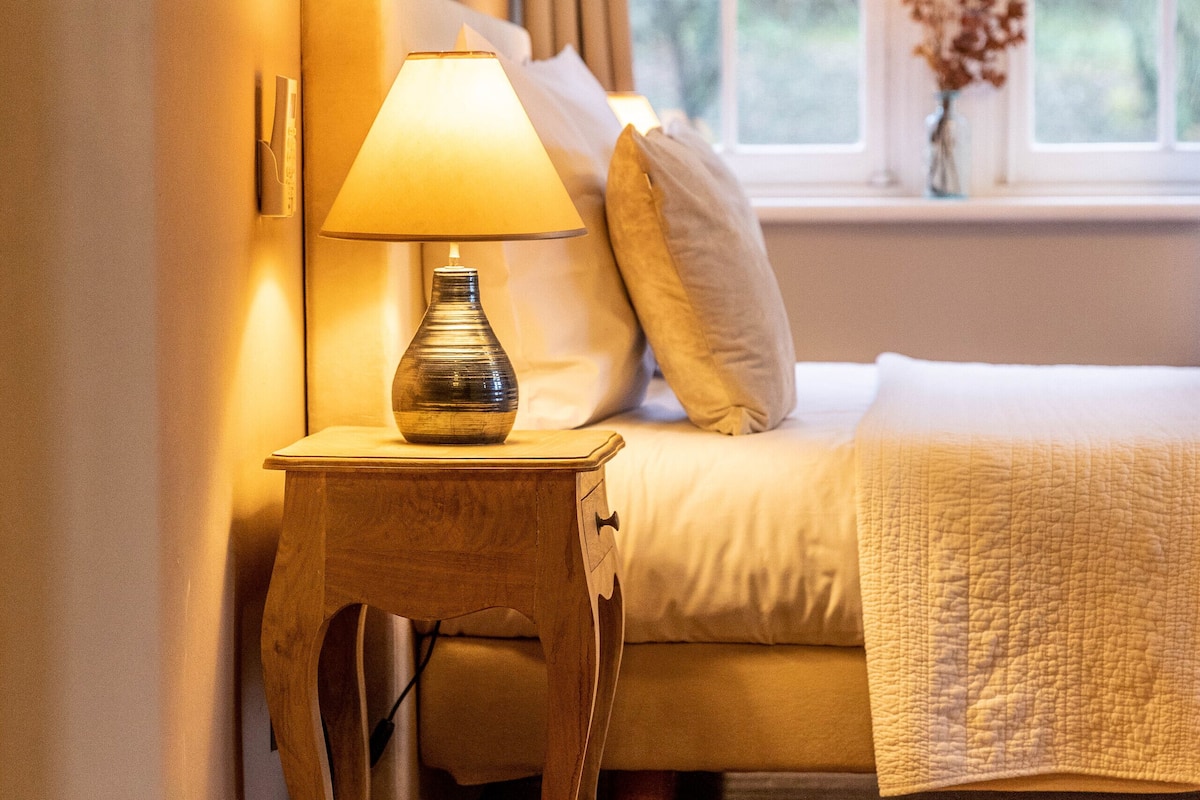 Un dormitorio con una mesita de noche de madera, una lámpara, una cama con colcha y una ventana con cortinas.