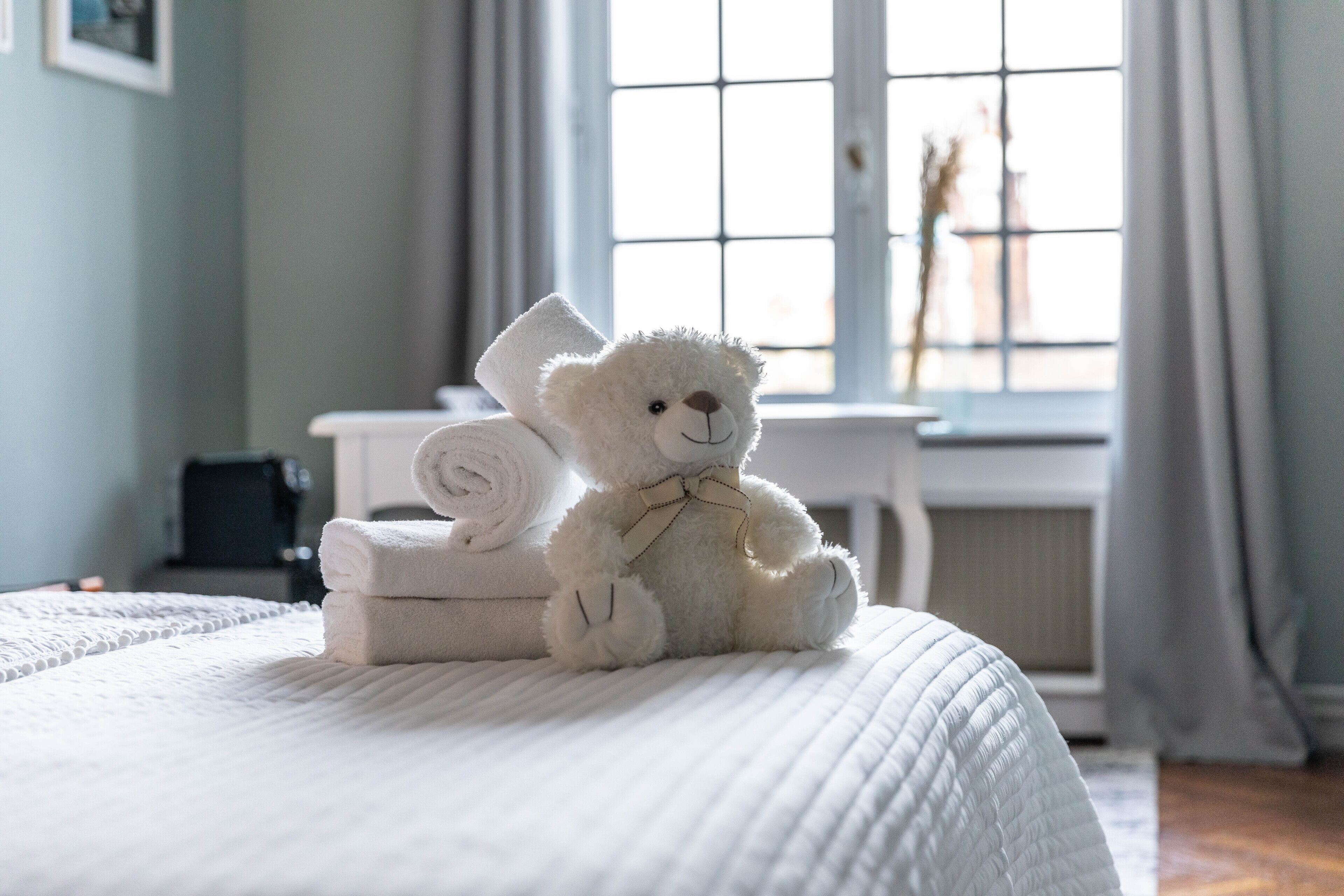 A bed with a white quilt, a white teddy bear, and rolled towels on it.