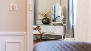 A room with a white vanity, a mirror, a vase with dried plants, and a wooden chair. - Ô Château (Hayange)