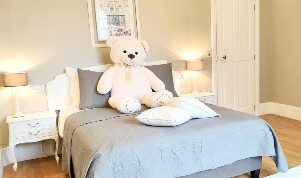 A bedroom with a large bed, a white teddy bear on the bed, two bedside lamps, a nightstand with drawers, and a door in the background.