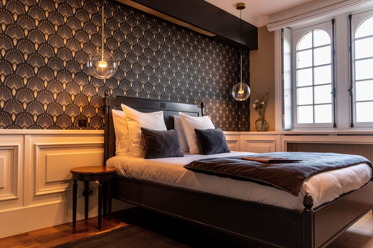 Un dormitorio con papel tapiz estampado, una cama con almohadas blancas y oscuras, un taburete de madera y un ventanal con marcos blancos.