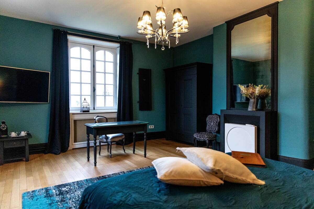 Un dormitorio con cama, escritorio, silla, televisor, espejo y ventana con cortinas.