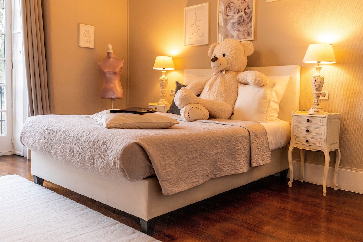 Una cama bien hecha con un gran osito de peluche, dos lámparas de noche y un aparador con cajones.