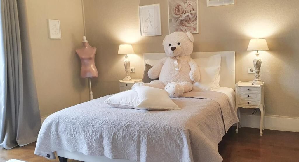 Et soveværelse med en stor seng, to sengeborde med lamper, en bamse, en mannequin og indrammede kunstværker på væggen.
