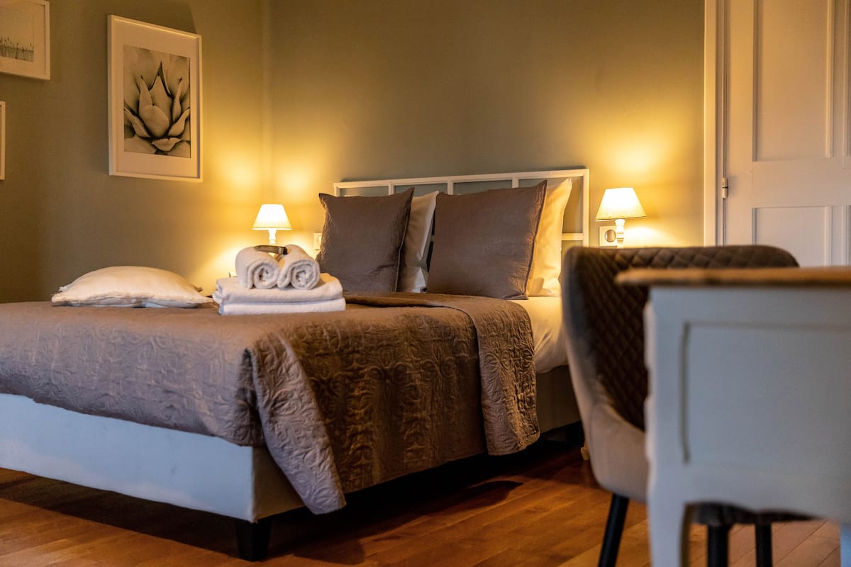 Una cama bien hecha con almohadas, una lámpara de noche y una toalla enrollada sobre la mesita de noche.