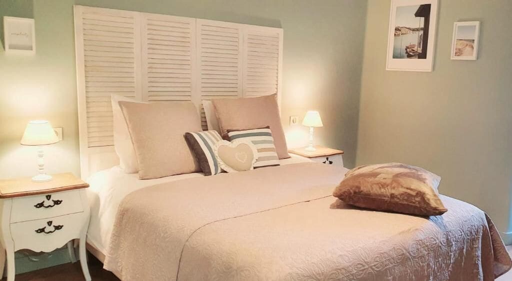 A neatly made bed with a beige bedspread and several pillows, two bedside tables with lamps, and a wall with framed pictures.
