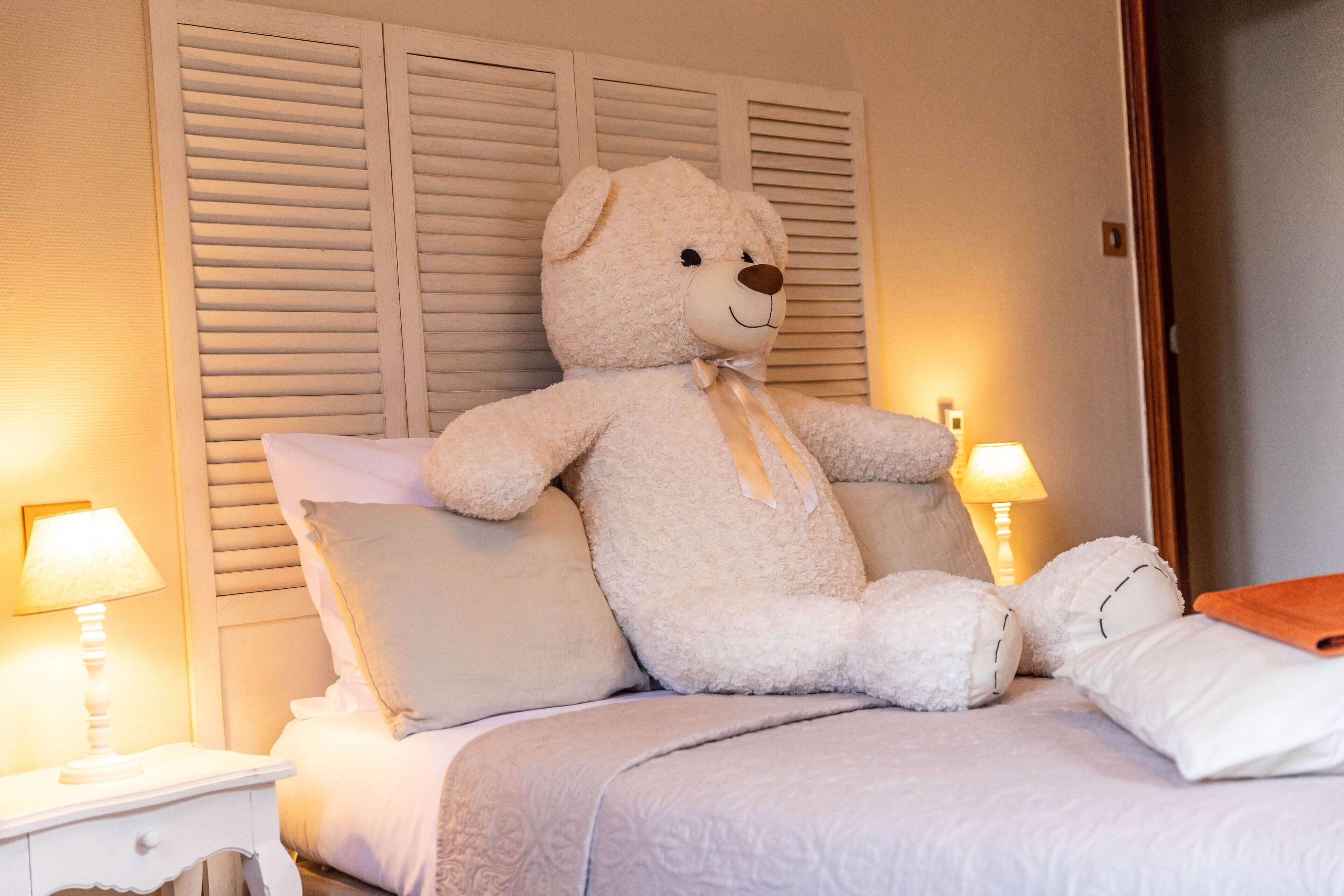 Ein Bett mit einem großen weißen Teddybären, weißen Kissen und einem Nachttisch mit einer Lampe.