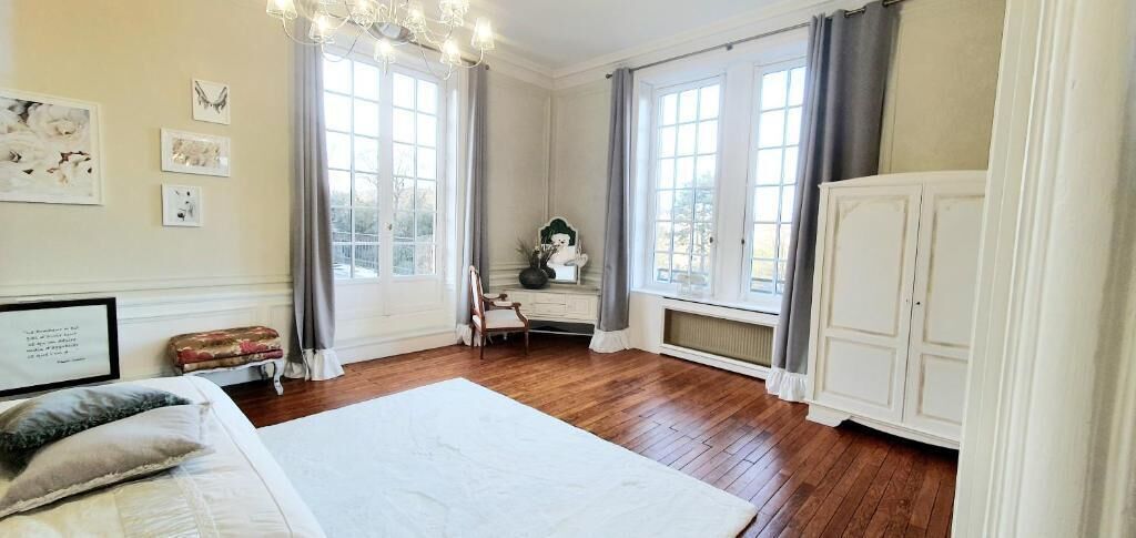 Ein Schlafzimmer mit einem großen Bett, einem Schminktisch, einem Stuhl und einem Schreibtisch. Zwei Fenster mit Vorhängen, eine Kristallleuchter und Kunstwerke an den Wänden sind zu sehen.
