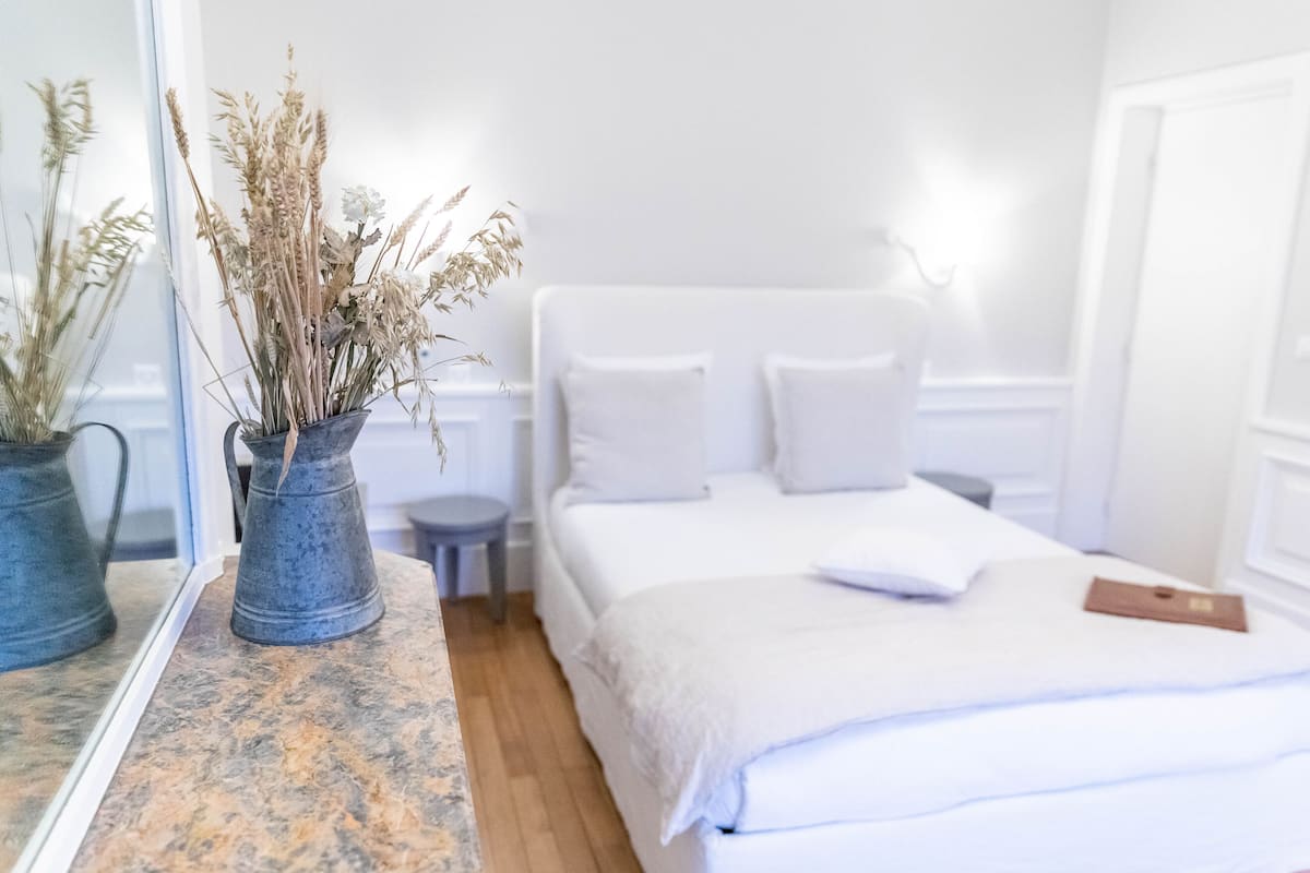 Un dormitorio con una cama, una mesita de noche, un espejo y dos jarrones con plantas secas.