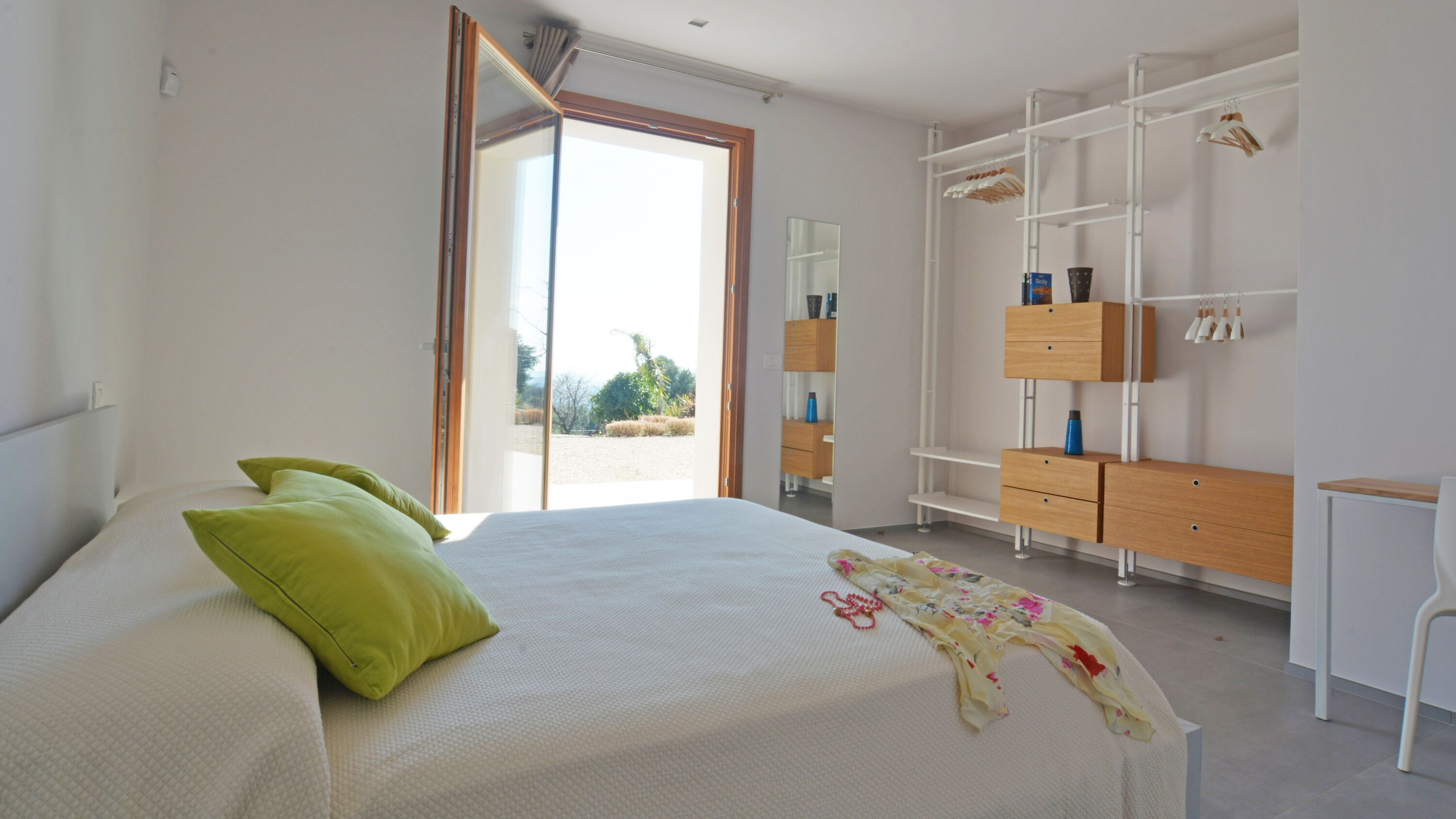 A bedroom with a bed, green pillows, a wooden dresser, a white chair, and a large window.