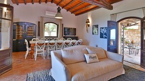A cozy living room with a sofa, dining table, and chairs. There is a TV, a window with curtains, and a stone wall outside. - Villa Passulunara 14 (Collesano)