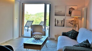A living room with a sofa, a coffee table, and a chair near a sliding glass door leading to a balcony with a chair and a table. - Villa Provenza 12 4 in Cefal (Cefalù)