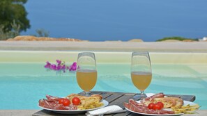 Mesa à beira da piscina com pratos de comida e dois copos de uma bebida amarela, com vista para uma praia.