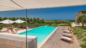 Ein Swimmingpool mit Liegestühlen und Sonnenschirmen, umgeben von viel Grün und freiem Blick auf das Meer.