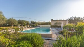 Ein rechteckiger Swimmingpool mit Liegestühlen und Sonnenschirmen, umgeben von viel Grün und einem Gebäude mit einem Ziegeldach.
