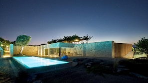 A modern outdoor pool area with a stone wall, a glass-enclosed lounge, and a wooden staircase.