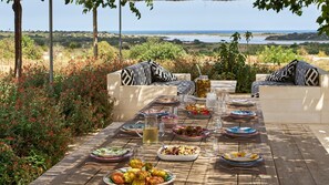 An outdoor dining area with a view of the sea, surrounded by greenery and furnished with zebra-patterned cushions and a wooden table set with various dishes and drinks.