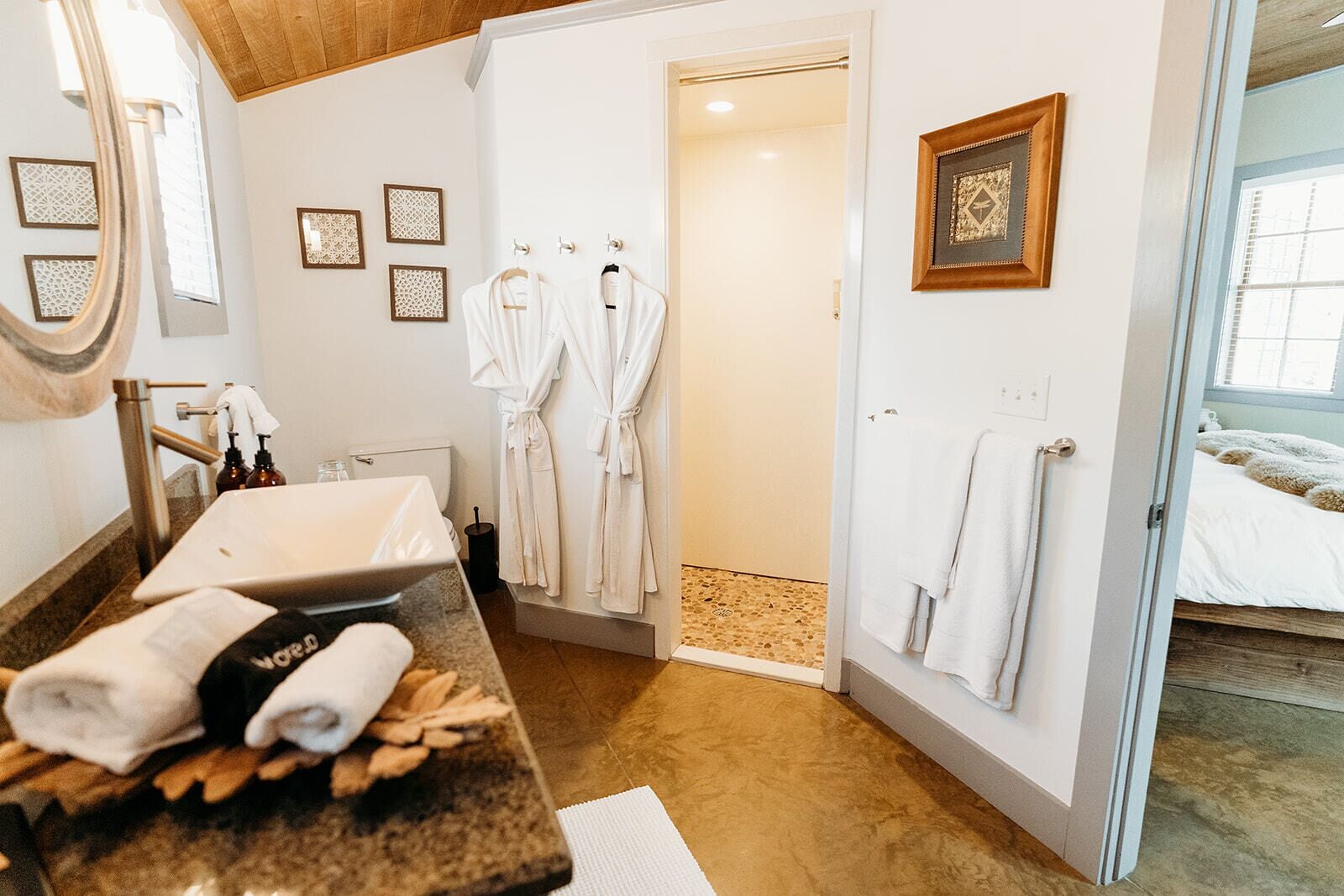 Un baño moderno con lavabo, espejo y toallero. Se observan dos albornoces blancos colgados en la pared.