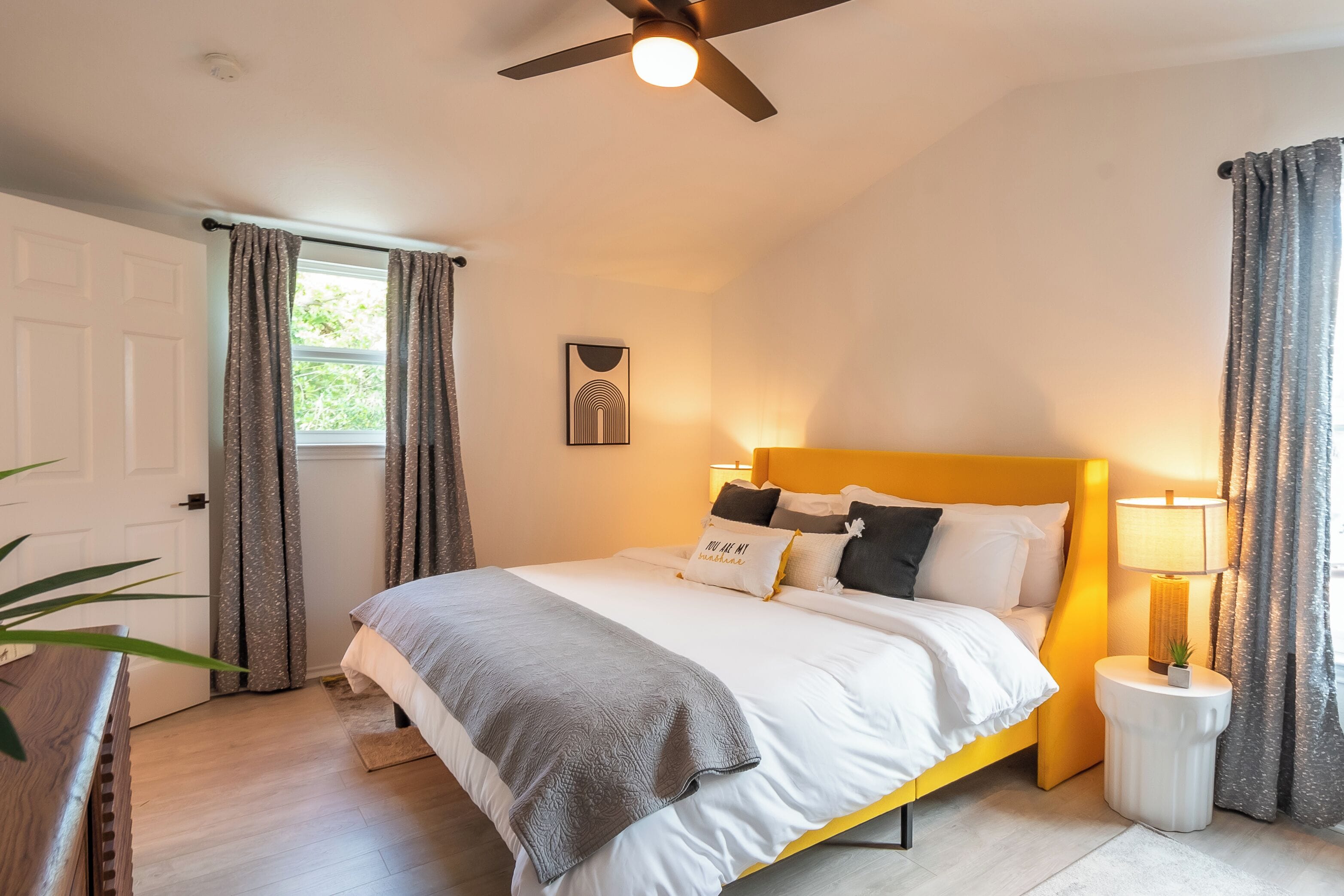 A bedroom with a bed, a ceiling fan, a bedside table with a lamp, and a window with curtains.