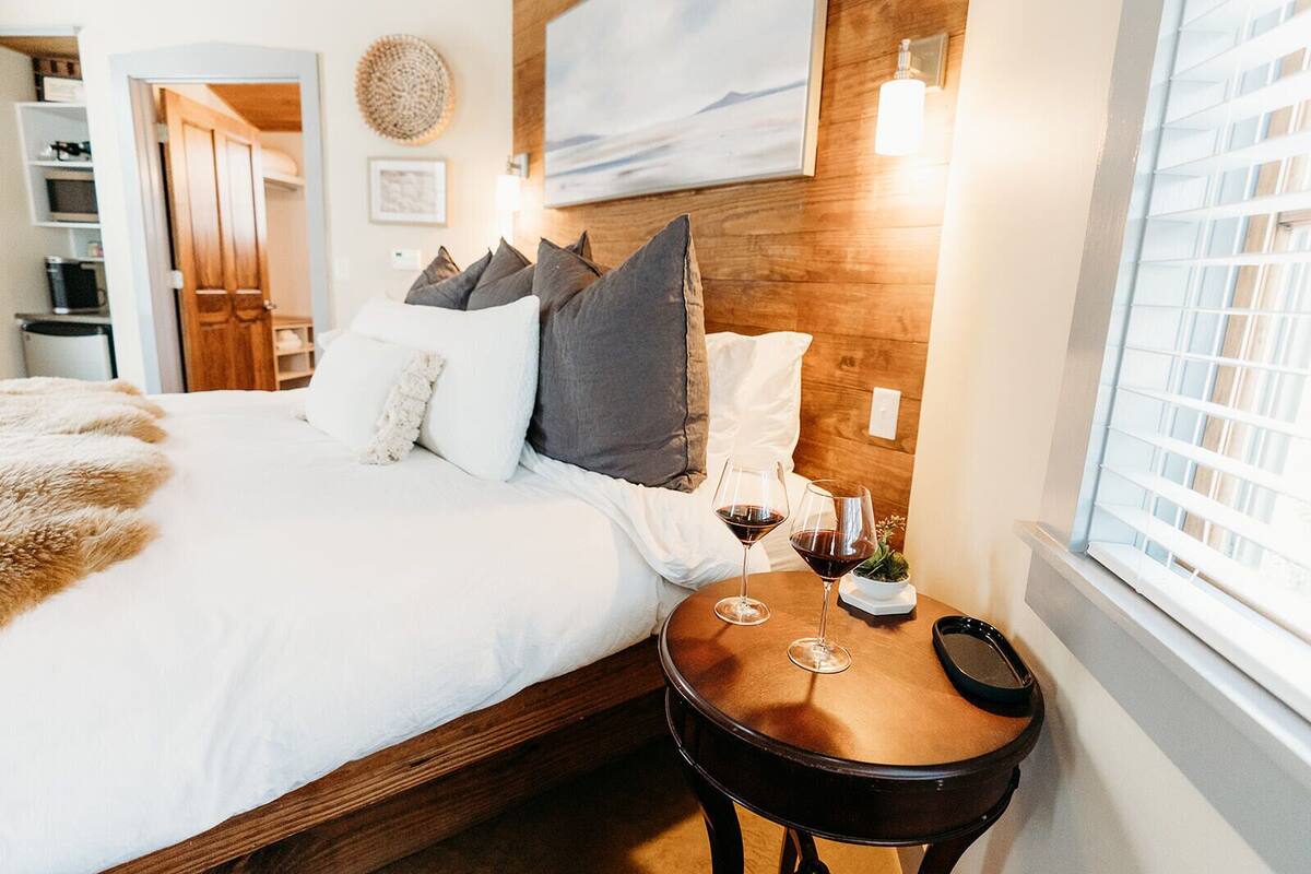 Una cama bien tendida con ropa de cama blanca y gris, un cabecero de madera y dos mesitas de noche con copas de vino.
