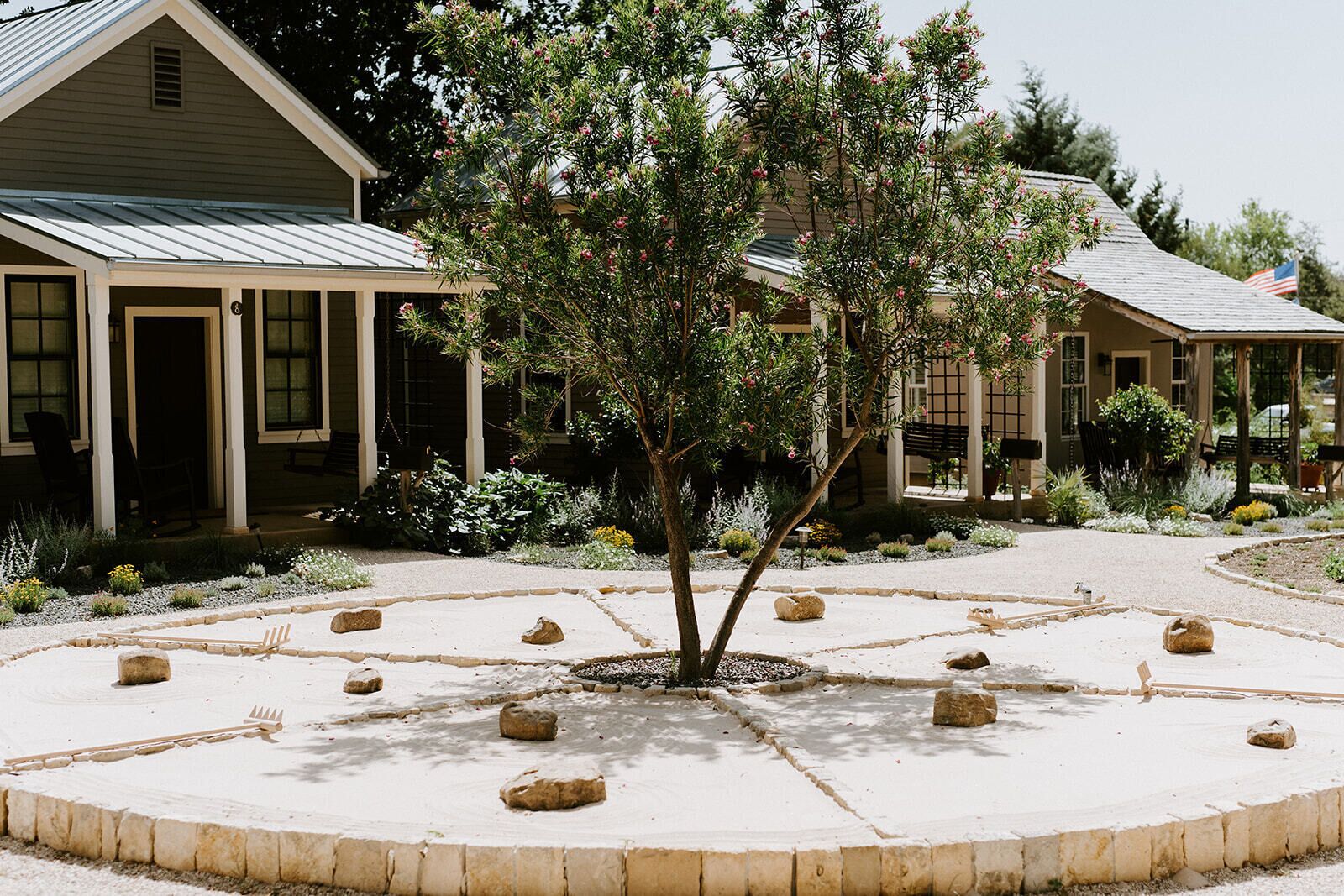 A building with a covered porch and a garden with a central tree and stone pathways.