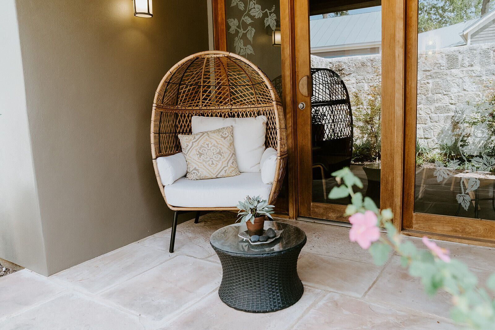 Una silla de mimbre con un cojín, una mesita con una planta y una puerta de vidrio que da a un muro de piedra y vegetación.