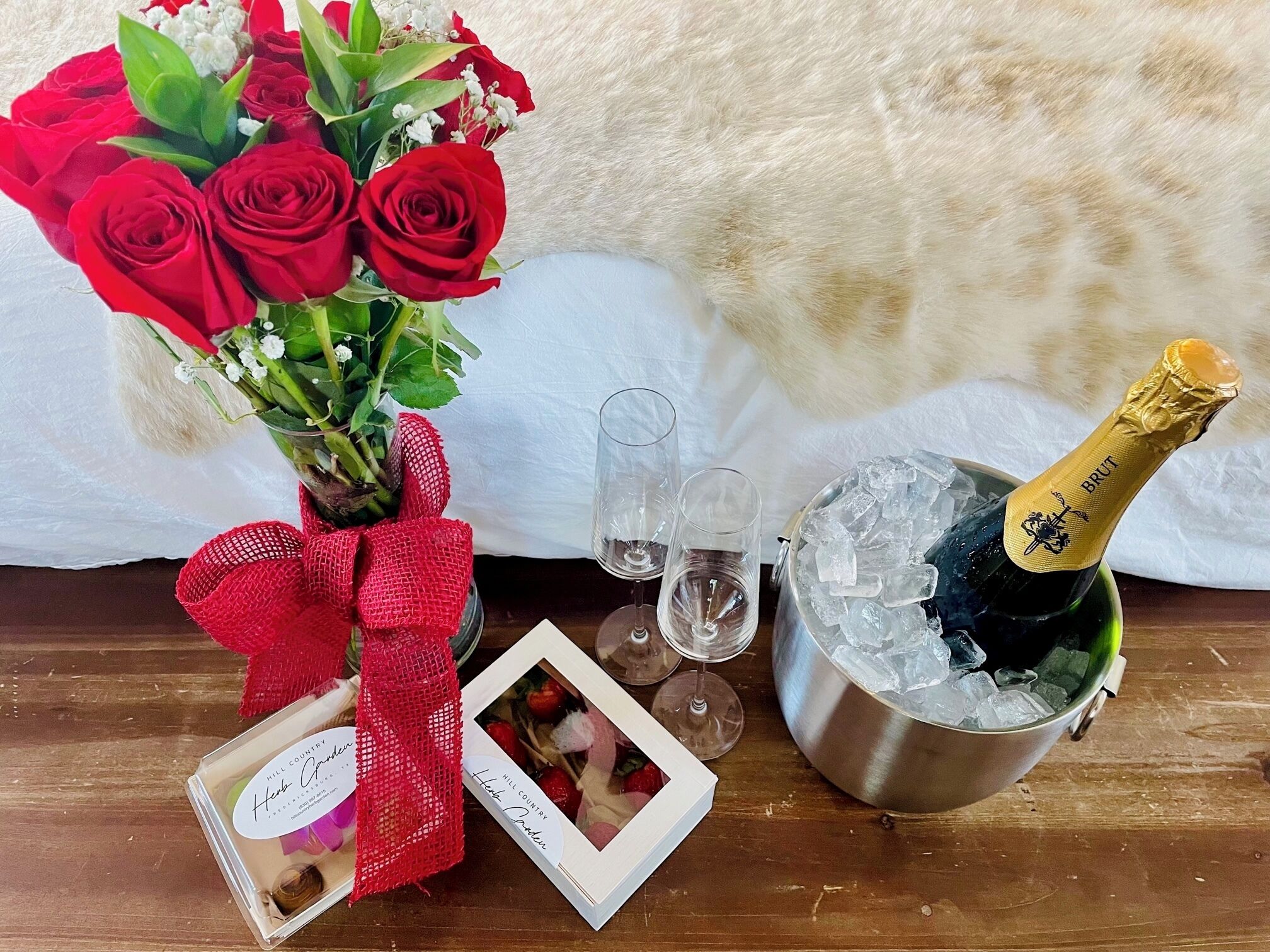 A bouquet of red roses, a bottle of champagne, ice in a bucket, two glasses, and a small framed photo on a wooden table.