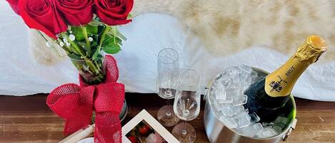 A bouquet of red roses, a bottle of champagne, ice in a bucket, two glasses, and a small framed photo on a wooden table.