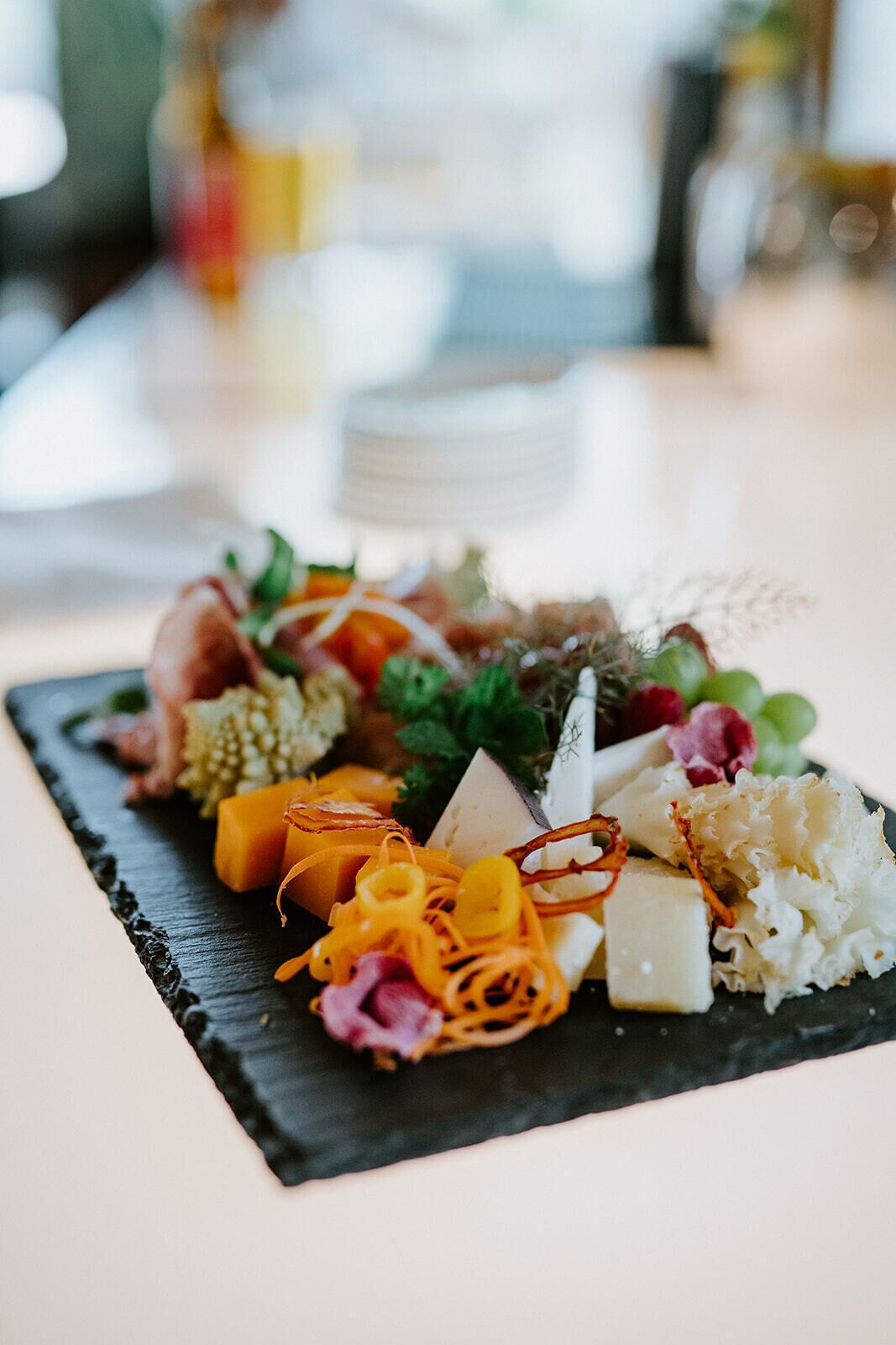 A black slate serving board with various cheeses, vegetables, and garnishes.