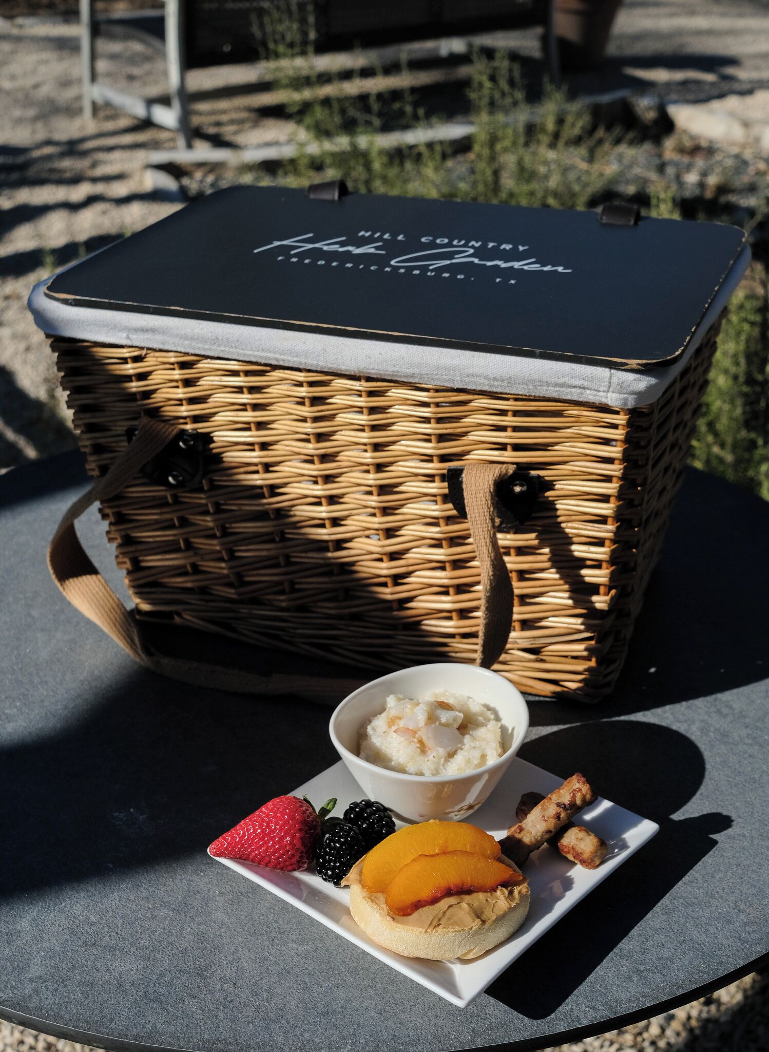 A wicker picnic basket with a lid, a bowl of dessert, and a plate with fruit and pastries.