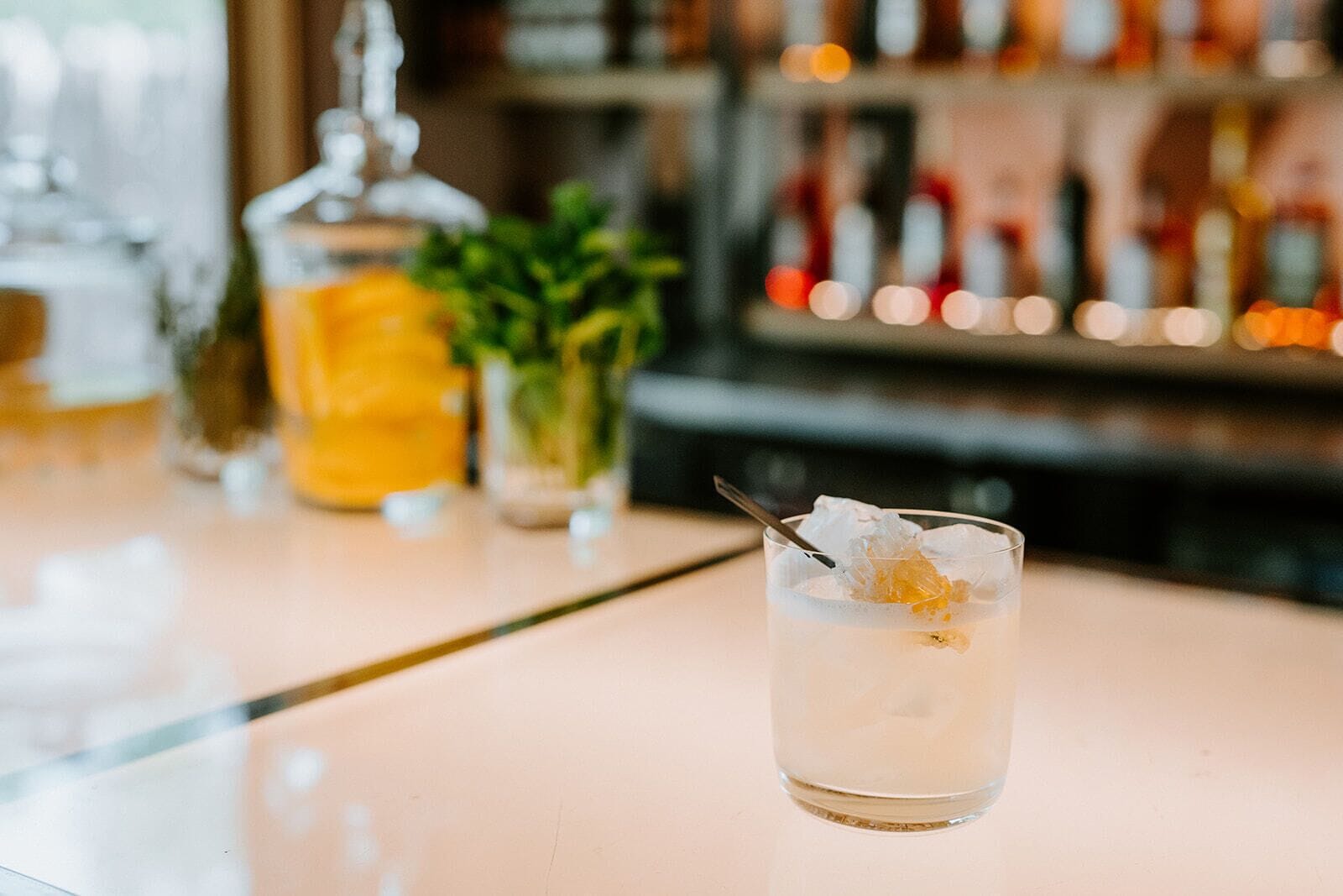 Un bar con un bicchiere trasparente pieno di bevanda, una bottiglia e una pianta in vaso sullo sfondo.
