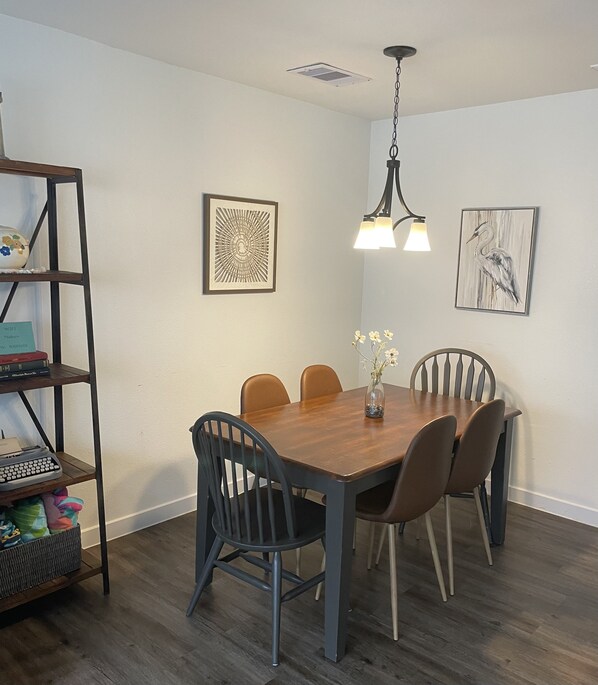 Een eetruimte met een houten tafel, zes stoelen, een hanglamp en twee kunstwerken in lijsten aan de muur.