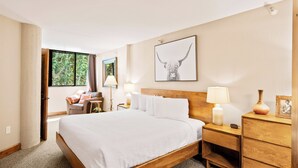 A hotel room with a large bed, a wooden nightstand, a desk, and a chair. There is a window with a view of trees, and a framed picture on the wall.