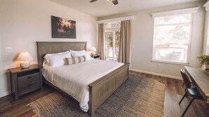 A bedroom with a wooden bed, two bedside tables with lamps, a ceiling fan, a large window with curtains, and a view of the outdoors.