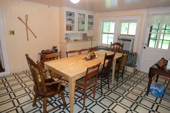 A dining room with a wooden table and chairs, a kitchen area with cabinets and a radiator, and a tiled floor. - Historic 1874 Farm House at Foot of Gunks (Wallkill)