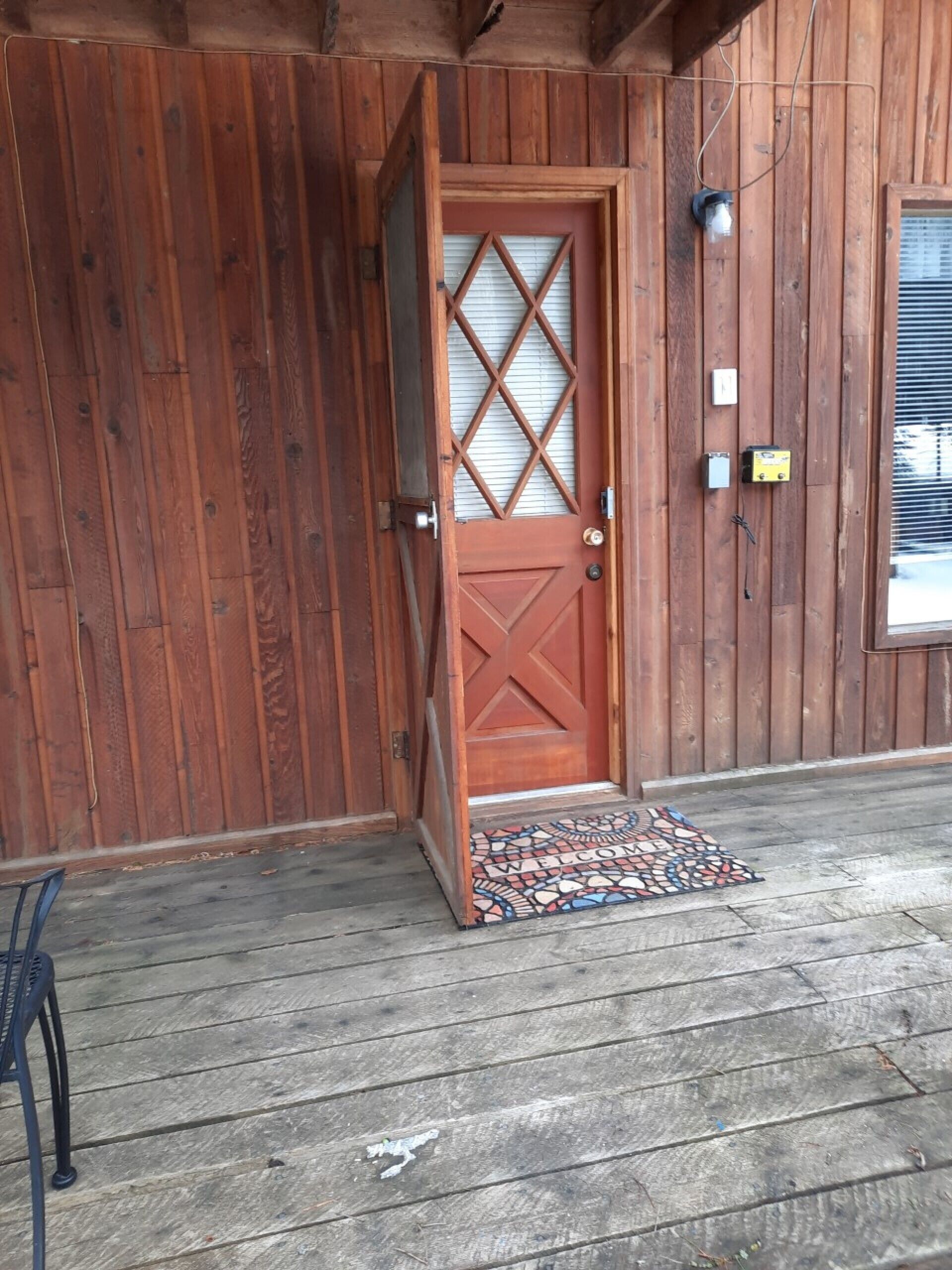 Une porte en bois ornée d’un panneau de verre à motifs losangés, un paillasson et une chaise en métal devant un bâtiment dont le revêtement extérieur est en bois.