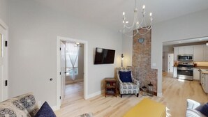 Uma sala de estar moderna com um sofá, uma poltrona estampada, um banquinho amarelo, uma TV fixada na parede e uma cozinha ao fundo.