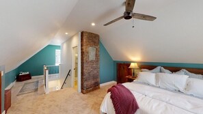 A bedroom with a sloped ceiling, a brick wall, a ceiling fan, a bed with white linens, and a nightstand with a lamp.