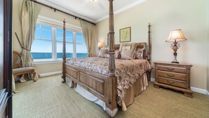 Ein Bett mit Baldachin, eine Blumendecke, ein Nachttisch mit Lampe und Blick auf den Ozean durch große Fenster.