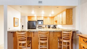 A modern kitchen with wooden cabinets, a granite countertop, a refrigerator, a microwave, and a toaster. There are three wooden bar stools. - Silver King 203 | 150 Yards to Lifts | Fireplace | Kitchen | End Unit | Mtn View | Pool & Hot Tub (Park City)