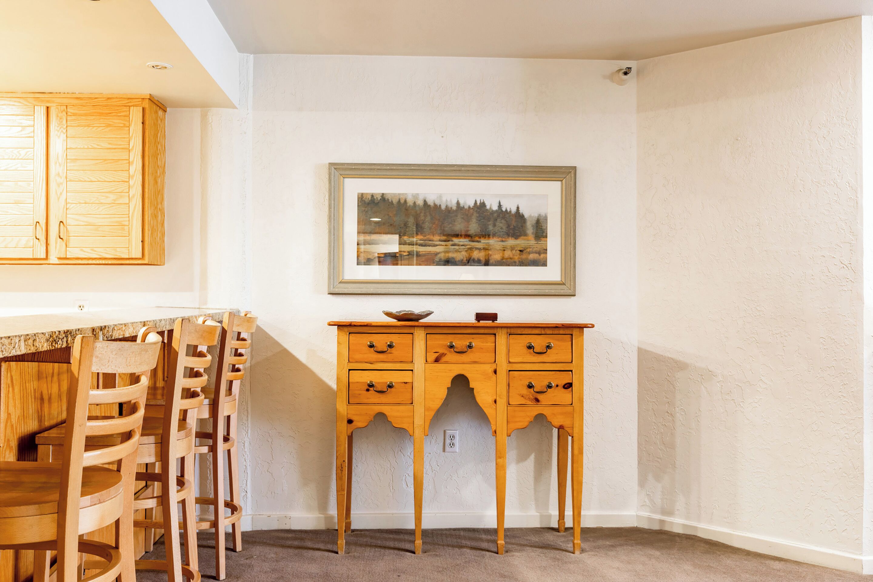 Un coin repas avec des chaises et une table en bois, un meuble en bois doté de tiroirs, et un tableau représentant un paysage, encadré, accroché au mur.